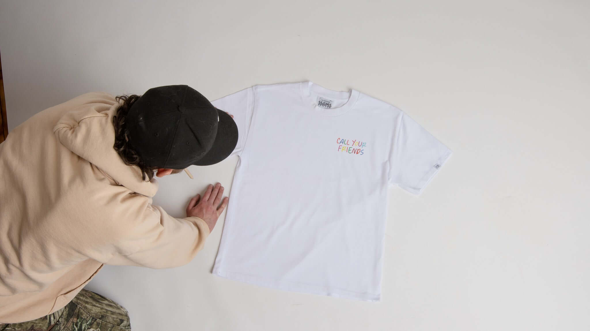 Person in a cap and hoodie laying out a white "Call Your Friends" Cereal Tee made of 100% cotton on a flat surface.